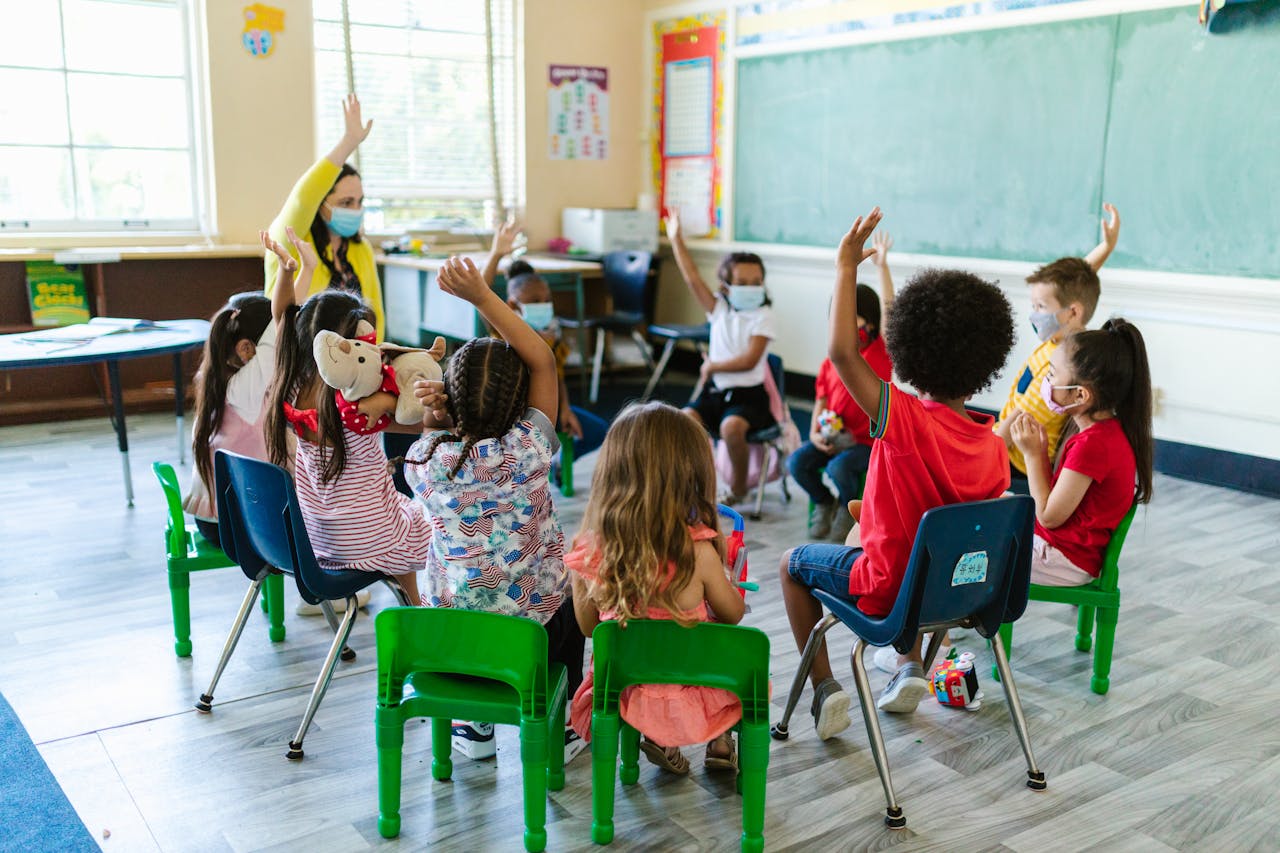 Home Diverse group of children in a classroom, raising hands, led by masked teacher. Bright and engaging learning environment.